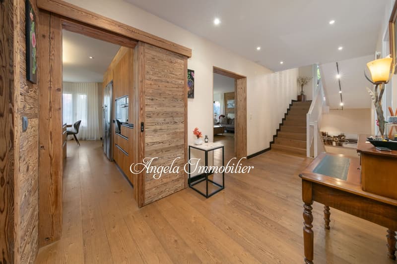 Spacious open-concept living area with hardwood floors, wooden accent walls, recessed lighting, and a fireplace. The room leads to a kitchen and staircase area.
