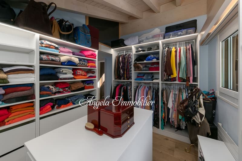 Spacious walk-in closet with built-in shelves and hanging rods, storing a variety of clothing items and accessories. The closet has a wooden dresser and a washing machine visible.