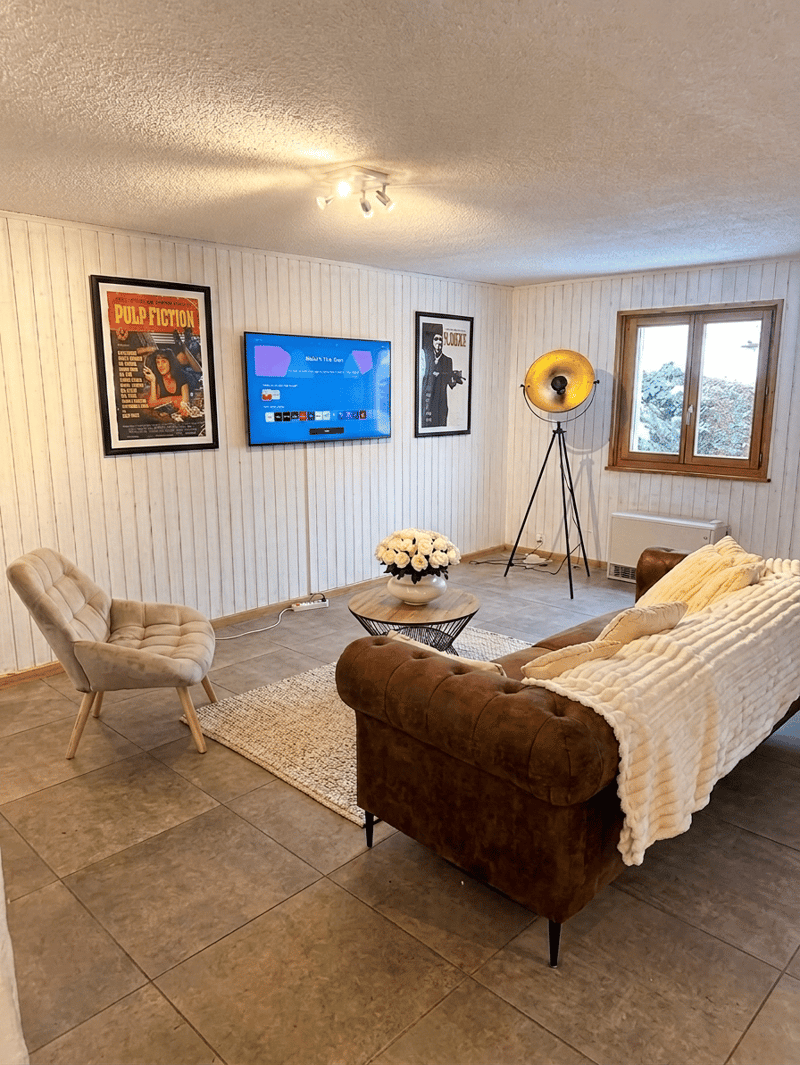 Living room with a brown sofa, a white chair, a coffee table with a flower vase, a TV mounted on the wall, a radiator, and a window.