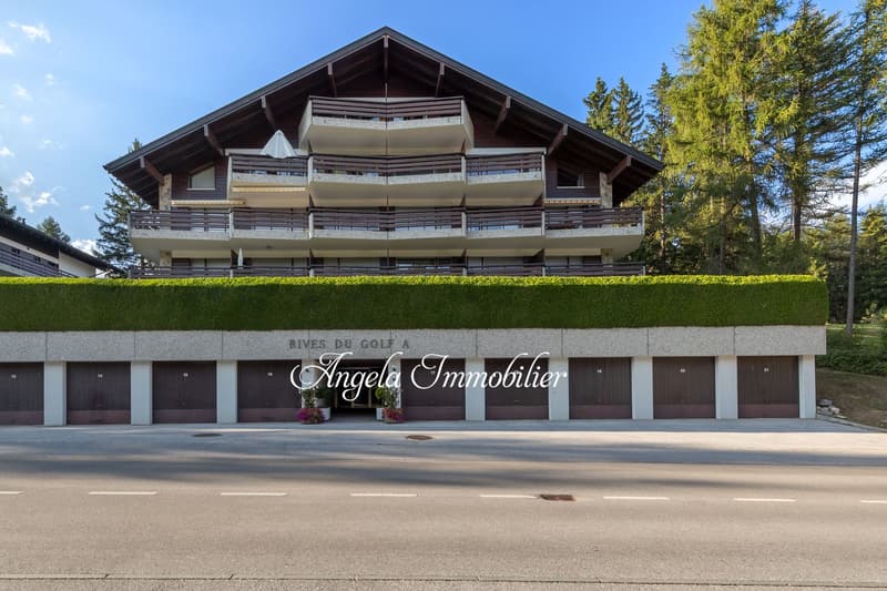Multi-story apartment building, modern design, balconies, brown wooden facade, green hedge, multiple garages, 'Rives du Golf A' sign, Angela Immobilier agency name, flowers by entrance, road in front