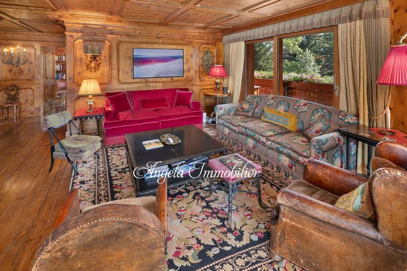 Cozy and rustic living room with wooden paneled walls and ceiling, large red velvet sofa, floral patterned armchairs, wooden coffee table, and a fireplace. The room has large windows overlooking a garden or outdoor area.