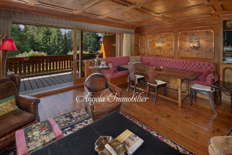 Spacious living room with wooden paneled walls and ceiling, large windows overlooking a balcony and outdoor scenery, plush pink tufted sofa, wooden furniture, and decorative wall sconces