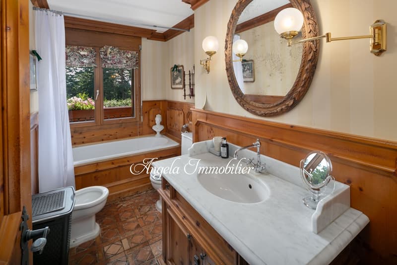 Spacious bathroom with wooden paneling, large round mirror, vanity with marble countertop, bathtub, and views of the outdoor garden through the windows