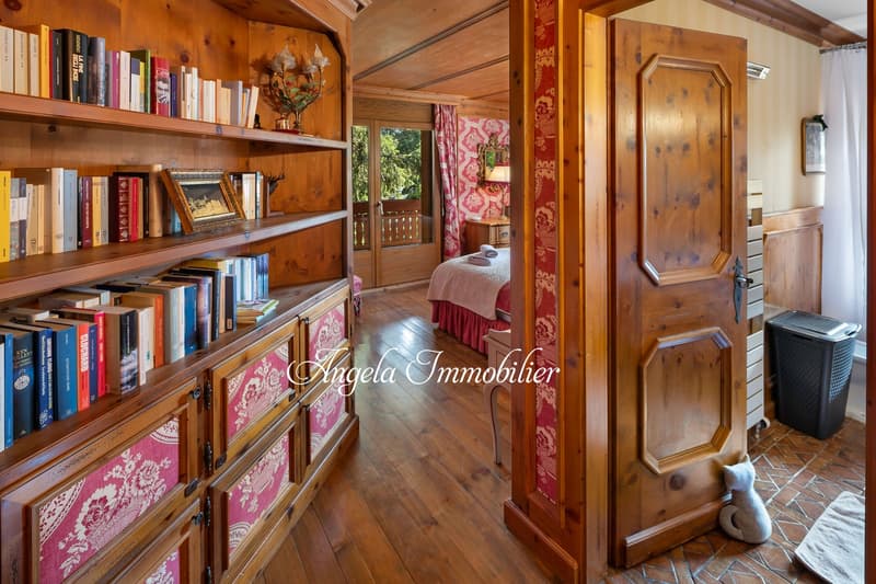 Cozy bedroom with wooden walls and shelves, pink patterned curtains, a bed with a bedskirt, and a fireplace visible in the background.