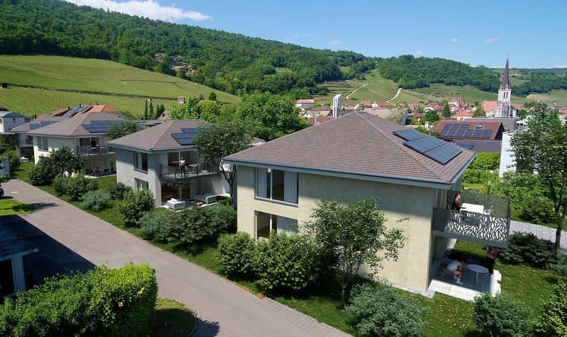Multi-story apartment building with balconies, solar panels on the roofs, surrounded by greenery and a small town in the background with a church steeple visible
