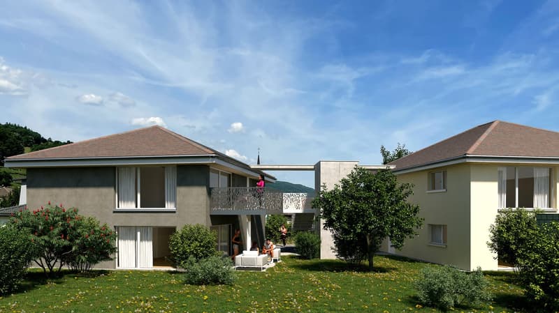The image shows a multi-unit residential building with a modern architectural design. The building has a mix of one and two-story units, with balconies or terraces visible on some of the units. The building is surrounded by a well-maintained garden with t