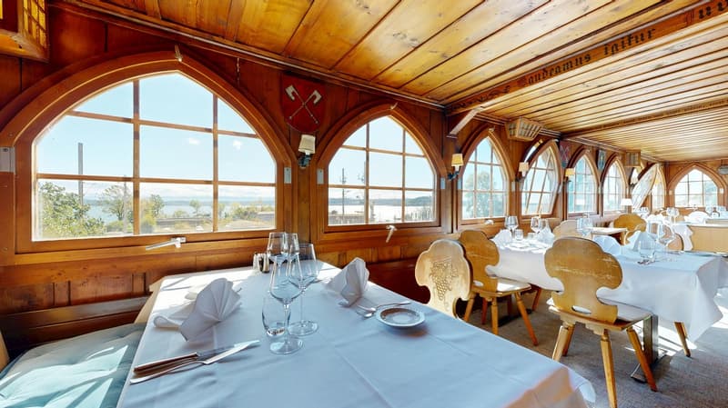 The restaurant has wooden walls and ceiling, arched windows with scenic views outside, and tables set up with white tablecloths, chairs, and wine glasses.