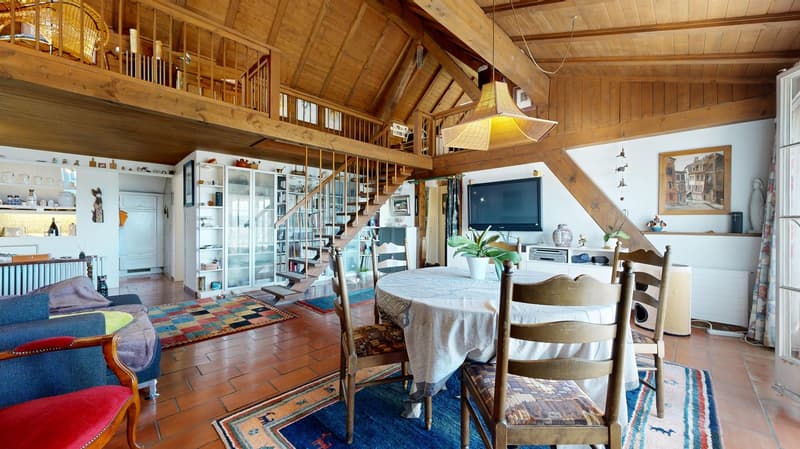The interior features a cozy dining area with a round table and chairs, a living area with a couch and armchair, and a kitchen with white cabinets and shelves. The room has a wooden floor and ceiling, with a staircase leading to a balcony or terrace. A te