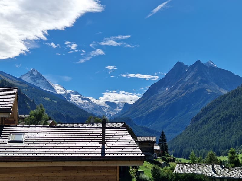 Building with wooden facade and tiled roof, located in a mountainous area, with a view of a snowy mountain range and a clear sky.