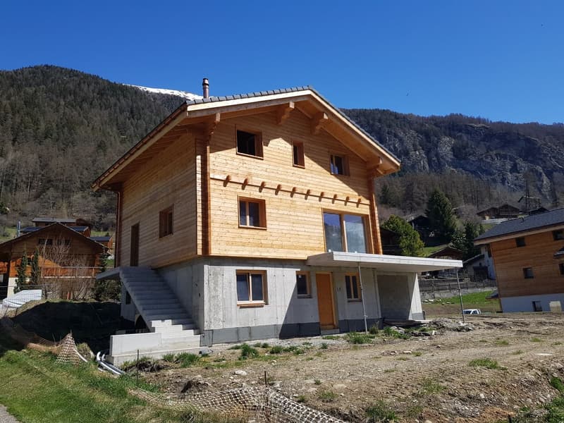 House with wooden facade, gray concrete ground floor, large windows, stairs, chimney, open terrace, mountainous backdrop.