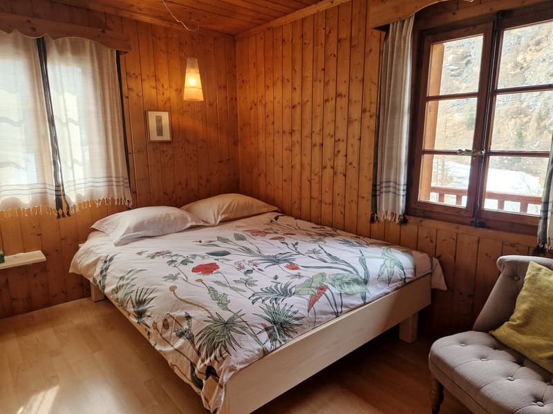 wooden walls, bed with floral cover, two pillows, small shelf, chair with pillow, two windows with curtains