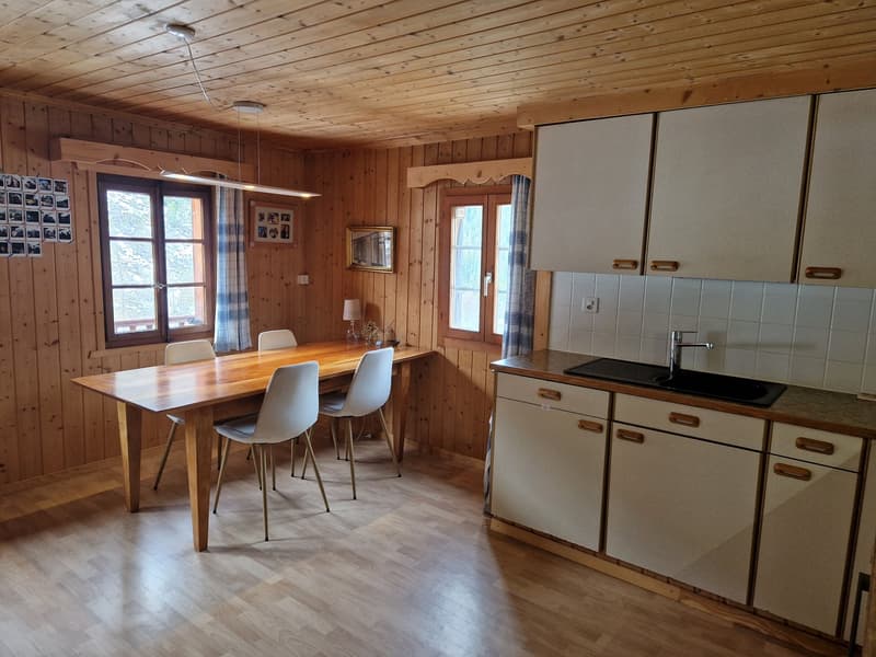 Wooden walls, wooden floor, dining table with chairs, kitchen with sink and cabinets, windows with curtains, picture frames on the wall