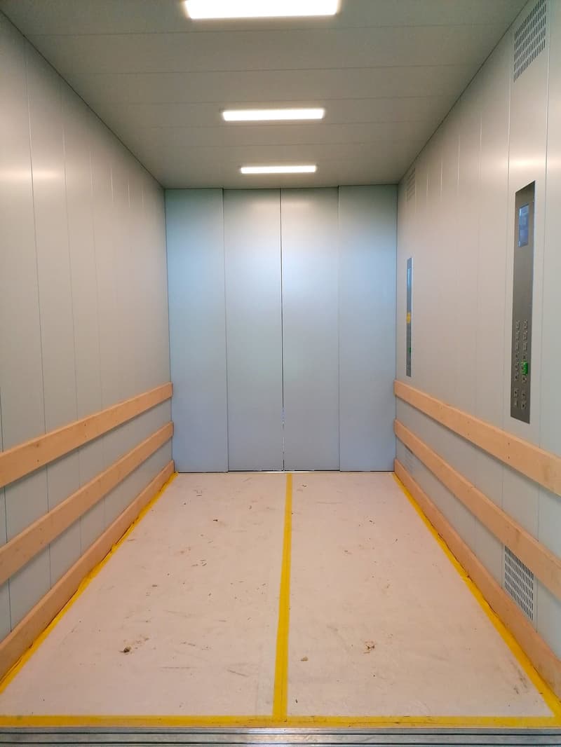 An empty elevator with gray walls, a white floor, yellow markings, and two doors.