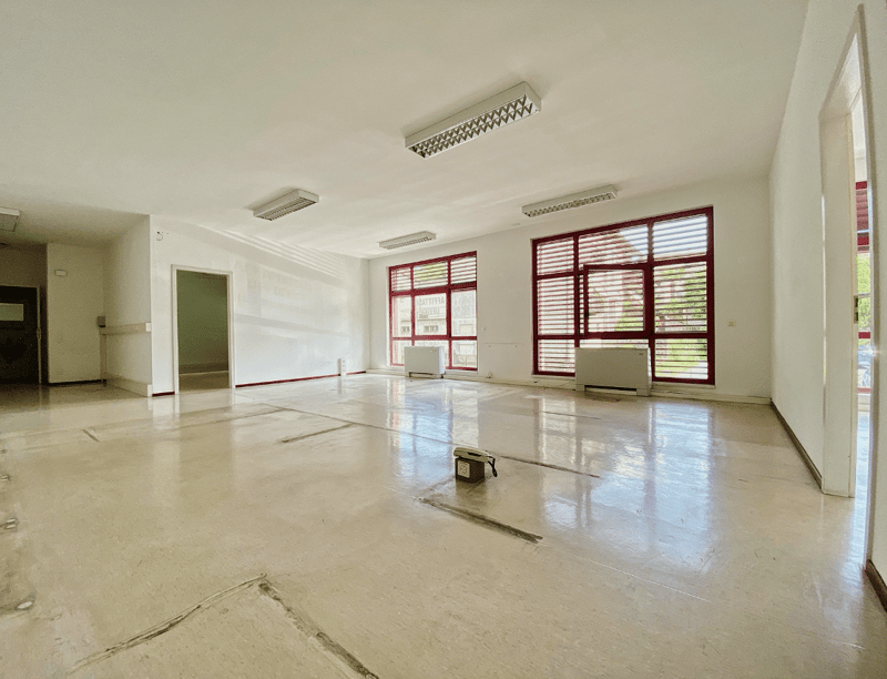 Empty room with white walls, concrete floor, and four large windows with red frames. Central heating units are located beneath the windows.