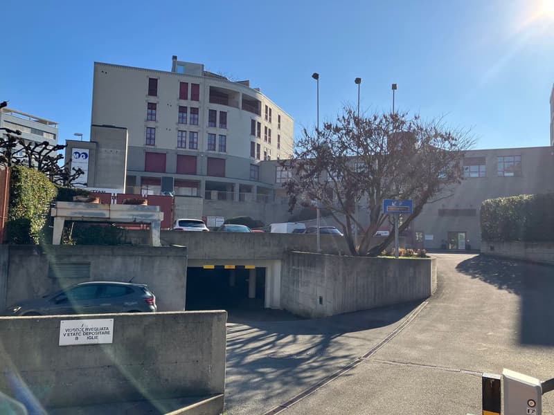 A multistory building with a parking lot in front, a garage entrance, a tree, several parked cars, and street lights
