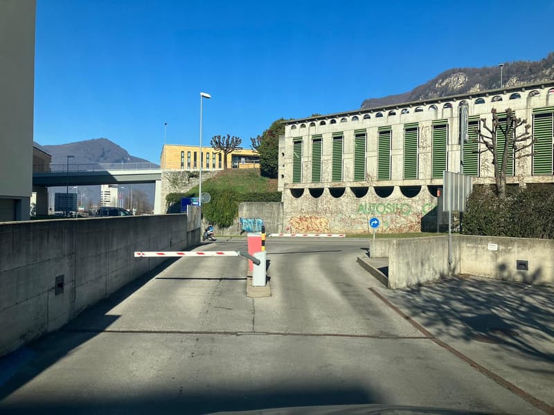Entrance to a parking facility with barriers, concrete walls, and buildings with graffiti in the background.