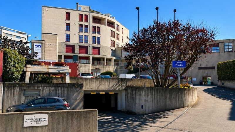 A multi-story building with red shutters, a curved facade, and a parking garage entrance. There are several cars parked in front of the building.