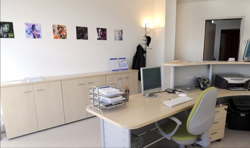 An office space with a desk, computer monitor, keyboard, mouse, and chair. There is a cabinet with books and papers, a coat rack with a coat, and a printer on the desk. A lamp is mounted on the wall, and there is a telephone on the desk. A basket with sci