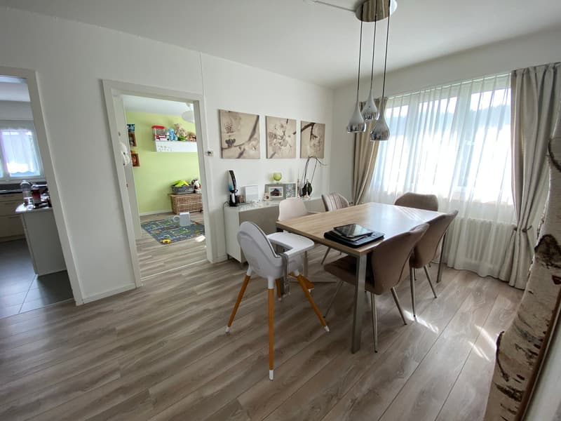 Dining room with wooden floor, white walls, dining table, chairs, white high chair, pendant lights, white curtains, rug, and a white cabinet with a basket on top.