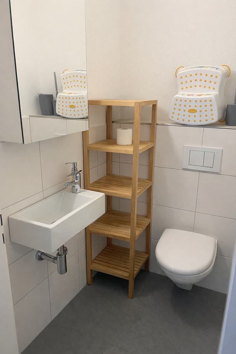 Bathroom with white tiles, wooden shelf, white sink, silver faucet, white toilet, mirror, and a roll of toilet paper on the shelf.