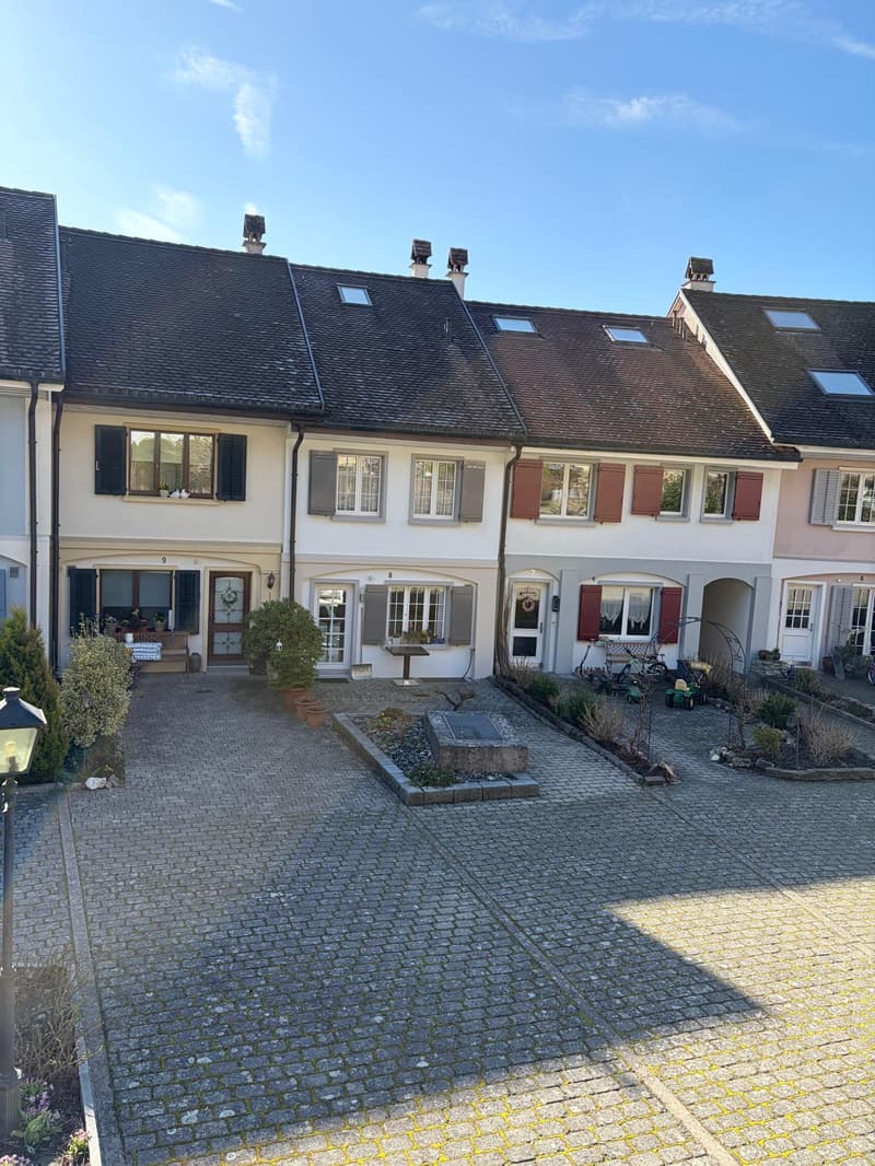 Row of terraced houses, brick walls, brown roofs, multiple windows, small garden in front, paved driveway