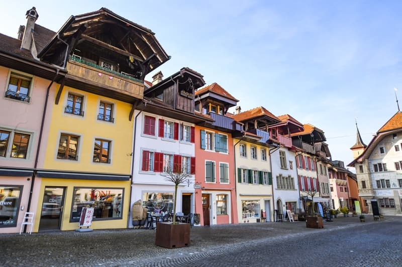 The image shows a quaint European town with traditional colorful buildings, some with wooden balconies and overhanging roofs. The buildings have a mix of architectural styles, with some having a more rustic, alpine appearance. There are shops and cafes vi