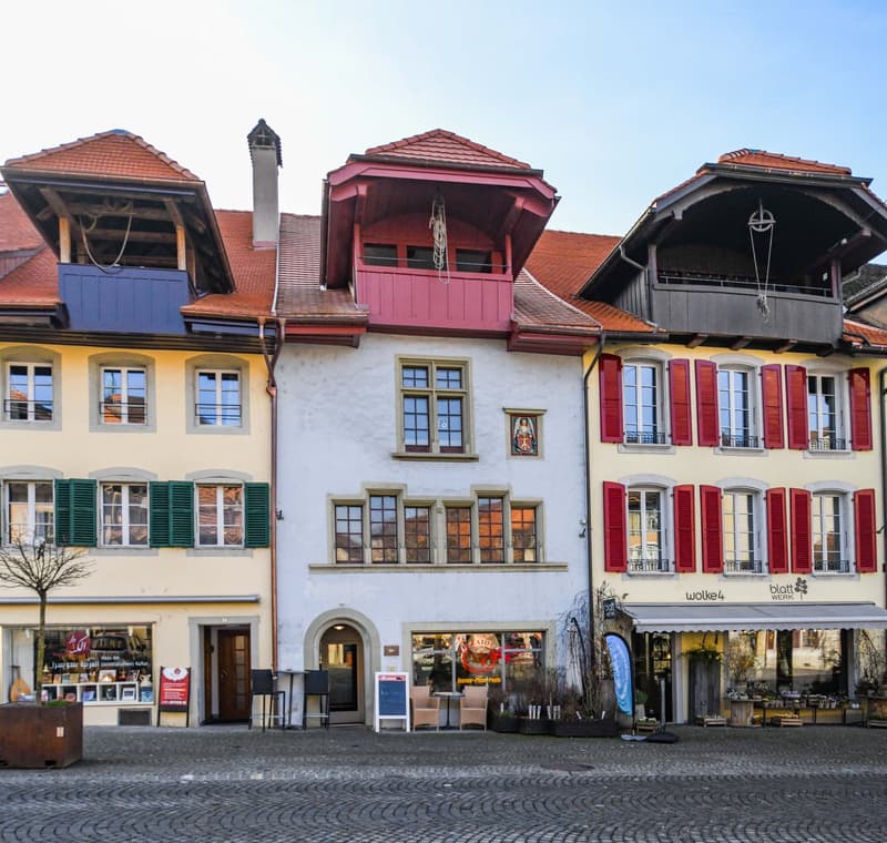 The image shows a row of traditional, colorful buildings with red tile roofs, wooden shutters, and decorative architectural elements. The buildings appear to be located in a town or city setting, with a cobblestone street in the foreground. The buildings