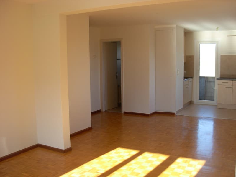 Empty room with wooden floor, white walls, open door, and a glass door leading to a kitchen.