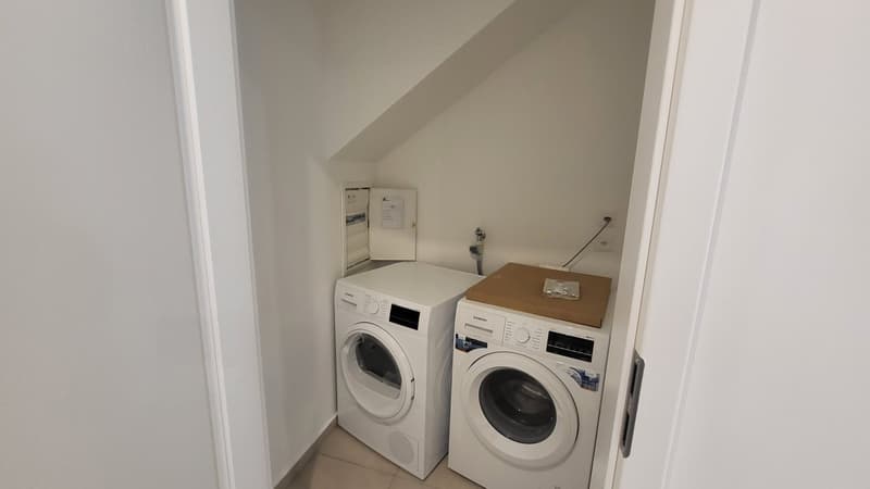 Small room with white walls, tiled floor, two white washing machines, one with a dryer on top, and a power outlet on the wall.