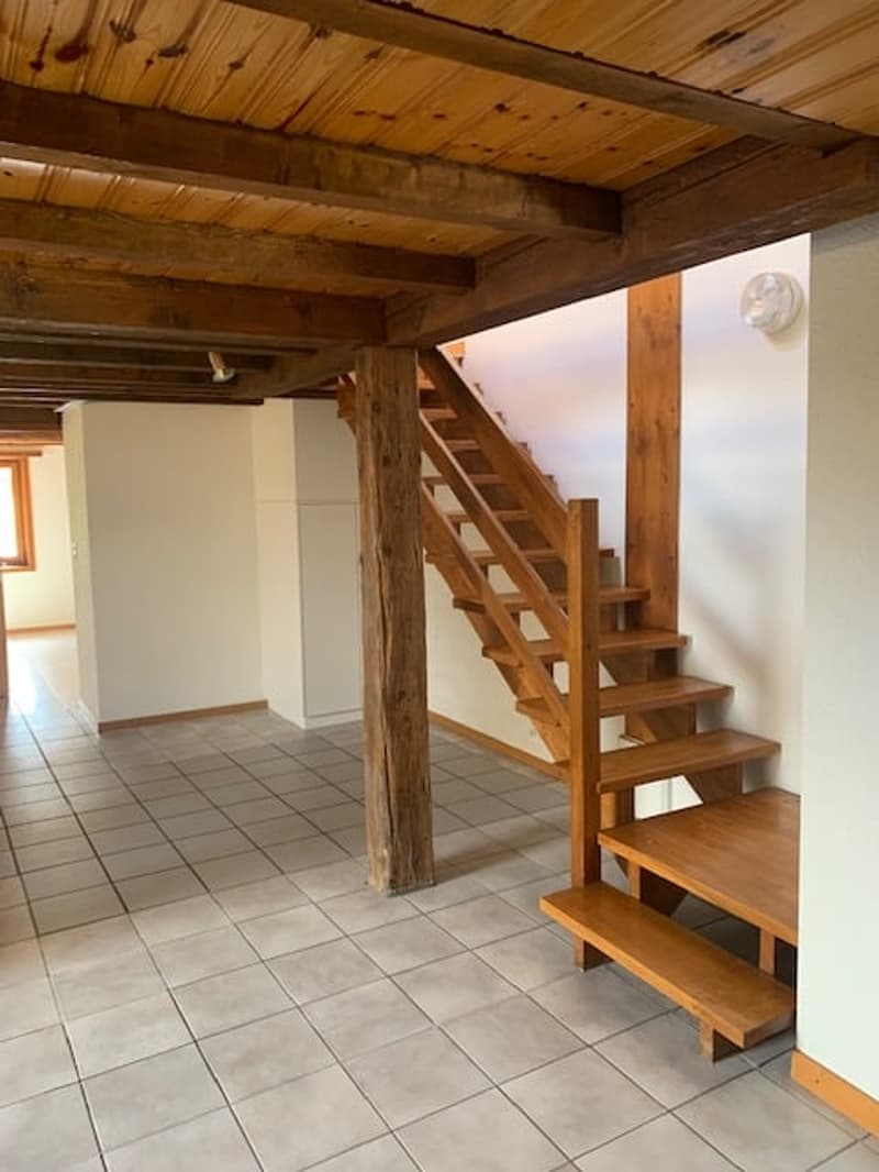 The image shows the interior of a rustic-style space with a wooden ceiling and exposed wooden beams. There is a tiled floor and a wooden staircase leading to an upper level. The space appears to be an open-plan layout with a clear separation between the d