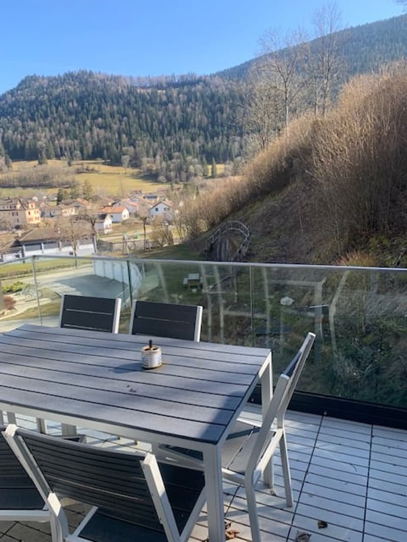 Outdoor deck with a table and chairs, overlooking a scenic mountain landscape with a village in the valley below.