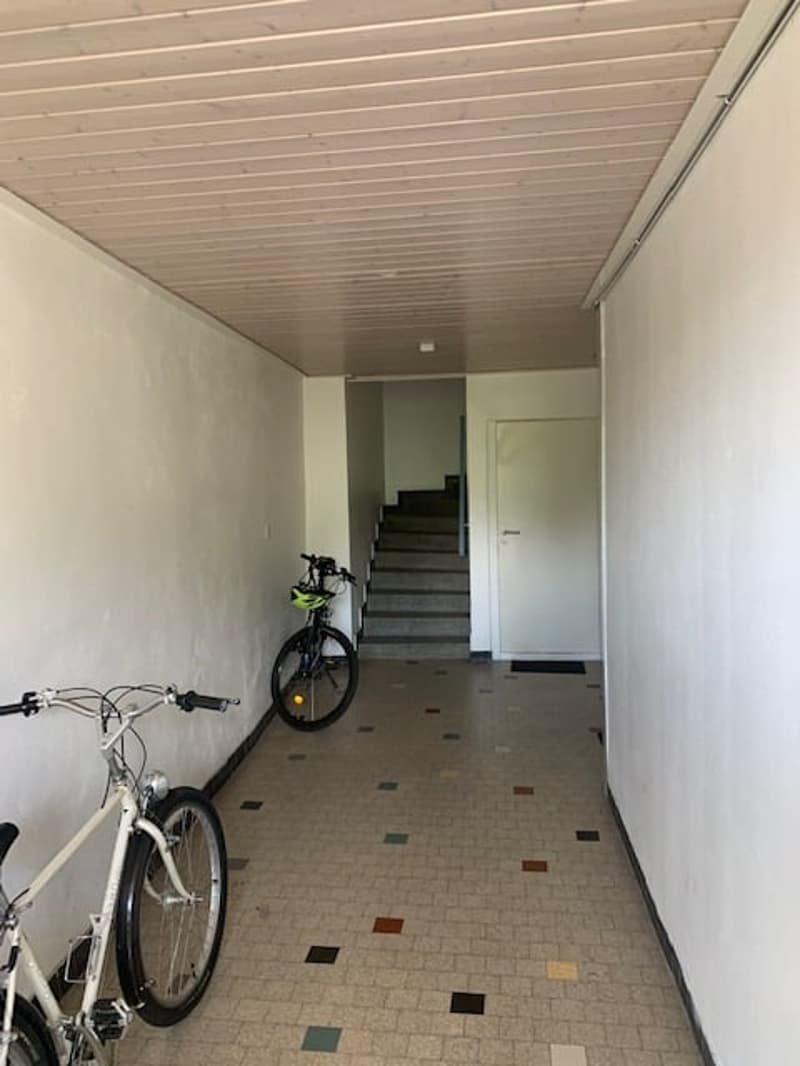 The image shows a hallway with a tiled floor and a wooden ceiling. There are two bicycles parked in the hallway, and a staircase leading up to the next floor is visible at the end of the hallway.