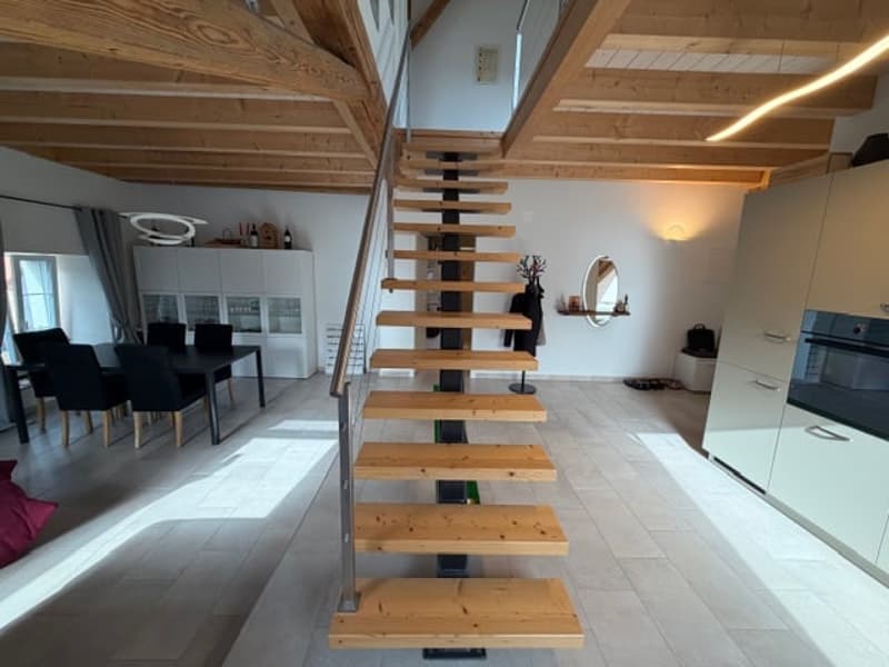 A modern house with wooden staircase, metal railings, and a kitchen area. The floor is tiled.