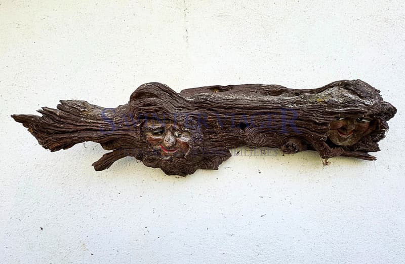/ a piece of art made from a tree trunk hanging on a white wall, with a face carved into the piece of wood