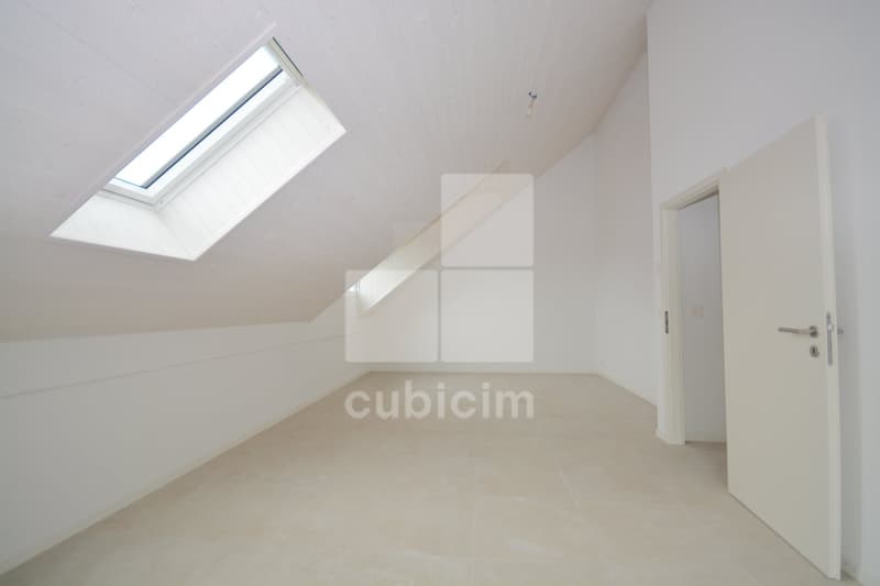 Attic room with slanted ceiling, large skylight window, white walls, and a white door leading to another room.