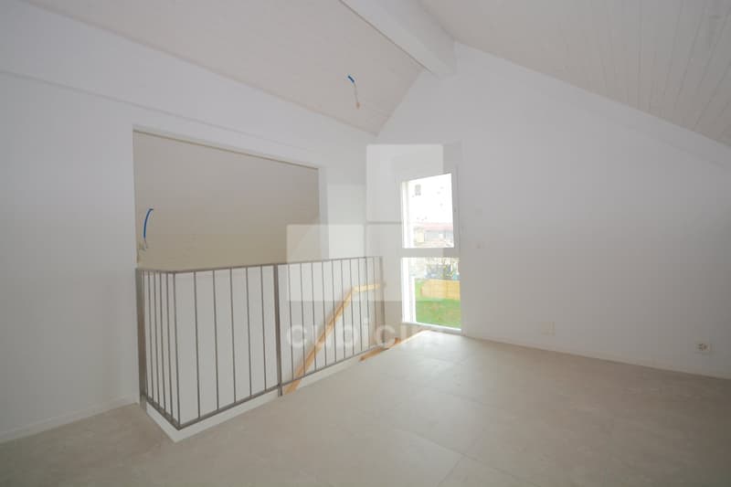 The image shows an empty room with white walls and a vaulted ceiling. There is a metal railing on the upper level, indicating a loft or second floor. The room has a large window that provides natural light. The floor appears to be tiled or made of a light