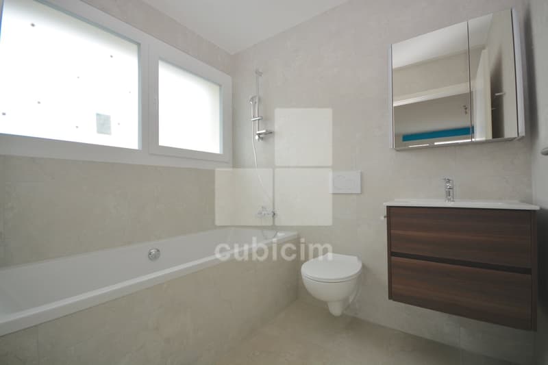 Spacious bathroom with large windows, white walls, tiled floor, bathtub, vanity cabinet with sink, and mirrored cabinet above the sink