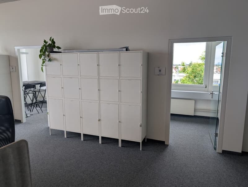 The image shows a room with a row of white lockers or storage units along one wall. There is a window overlooking some greenery outside, and the floor is covered in gray carpeting. The room appears to be an office or workspace setting.