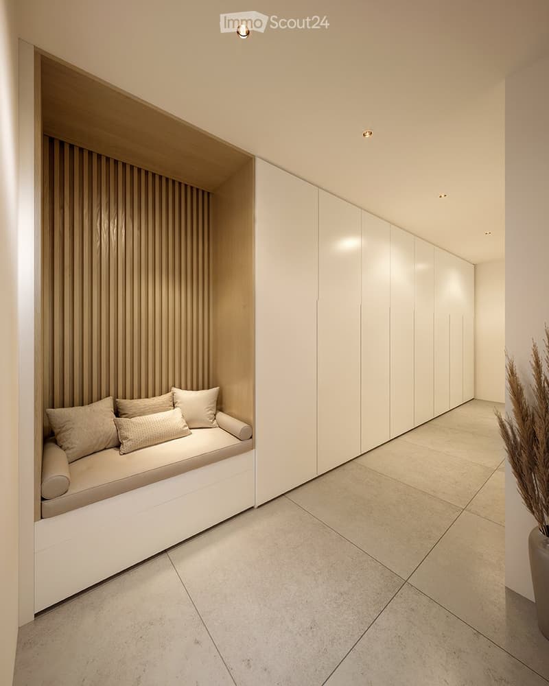 Spacious hallway with built-in storage cabinets, a cozy seating nook, and warm lighting. The floor is covered in light-colored tiles, creating a clean and minimalist aesthetic.
