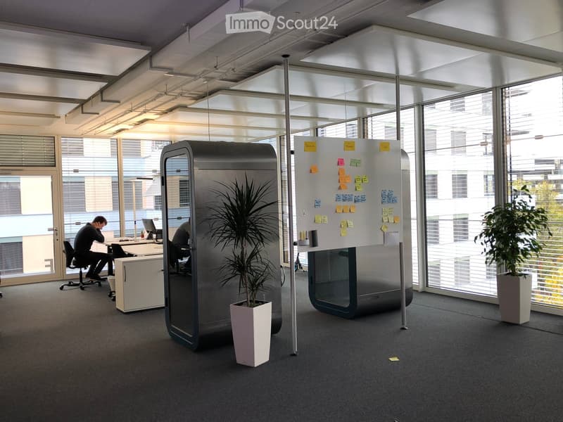 The image shows an open office space with large windows, a whiteboard with sticky notes, a refrigerator, and a potted plant. There are two people working at desks in the background.