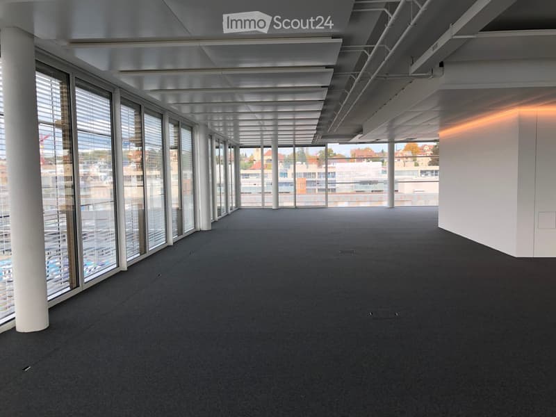Large open-plan office space with high ceilings, exposed beams, and floor-to-ceiling windows offering a panoramic view of the surrounding cityscape. The space features a grey carpeted floor and a clean, modern aesthetic.