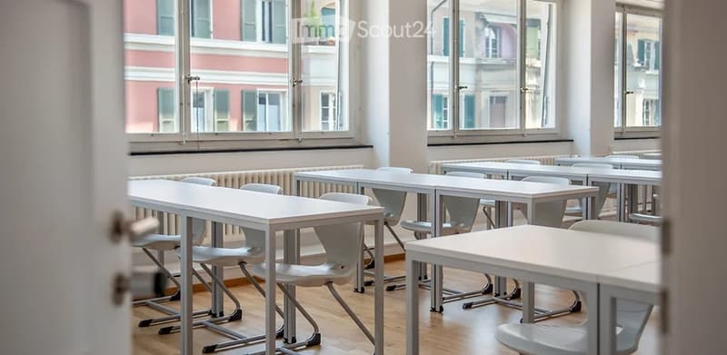 Empty classroom with tables and chairs arranged in rows, large windows facing the building.