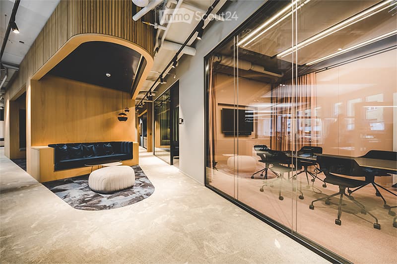 Meeting room with white walls, glass partitions, dark wooden sofa with cushions, glass table, chairs, ottoman, beige rug