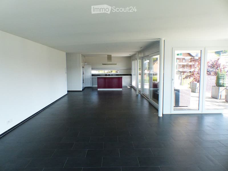 Empty room with white walls, black tile flooring, kitchen area with a red counter, a refrigerator and an extractor hood, sliding glass doors leading to a balcony with plants