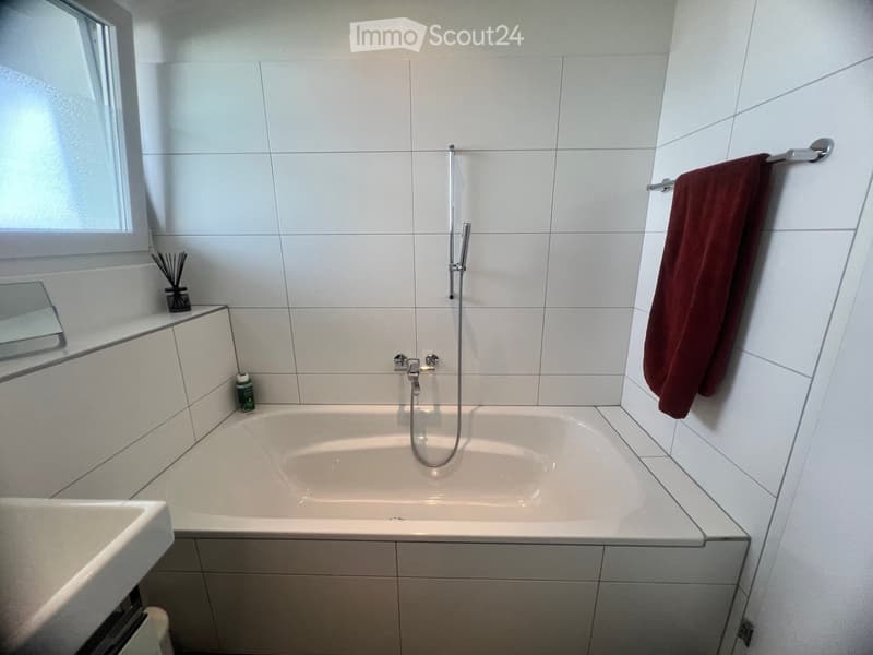 modern white tiled walls, large bathtub, separate sink, shower head, towel rack with red towel, mirror on the wall, green bottle on the counter, floor without visible flooring