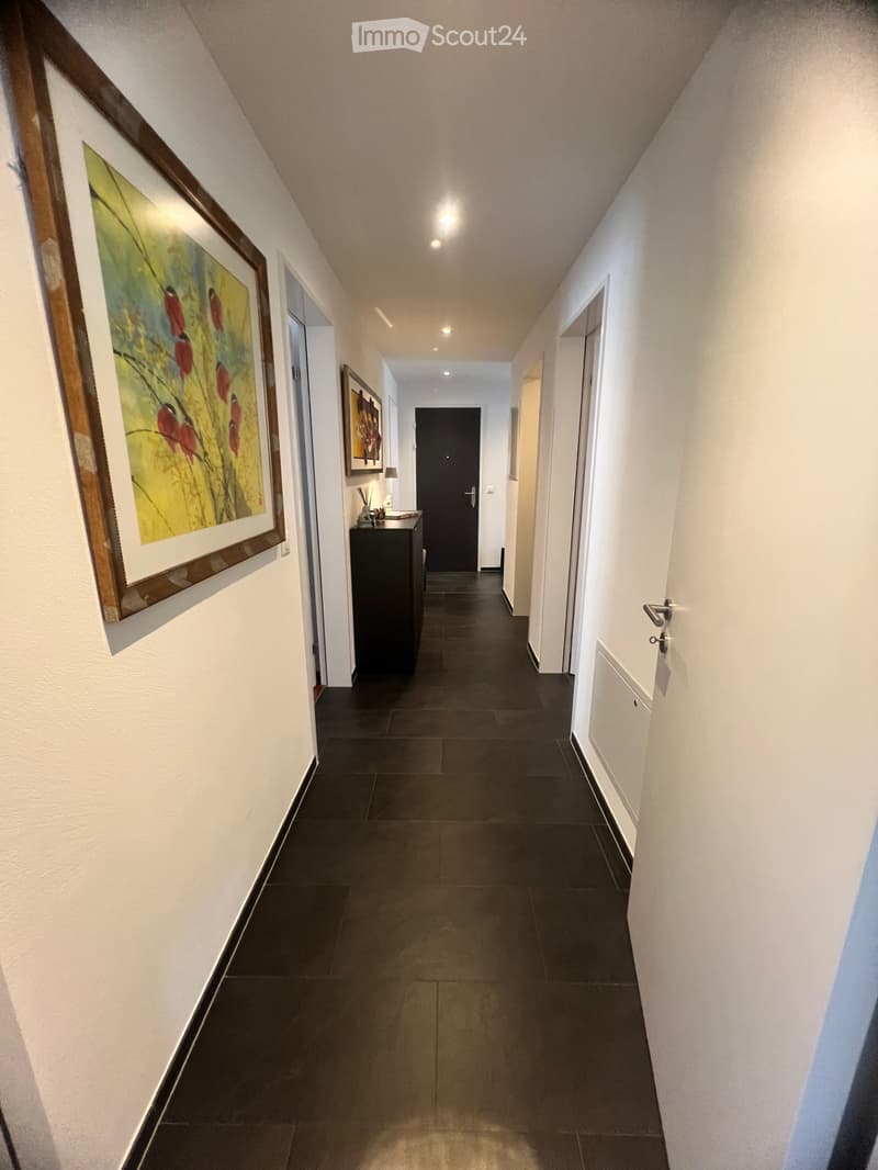 An apartment hallway with black tile flooring, white walls, and illuminated ceilings. The hallway is adorned with framed artwork on the wall and a black cabinet with a lamp in the distance. A few doors are situated on either side of the hallway.