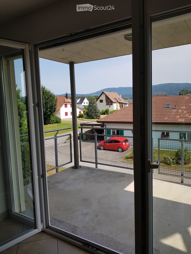 An exterior view through a sliding glass door onto a balcony with a metal railing and a red car parked in front of another house.