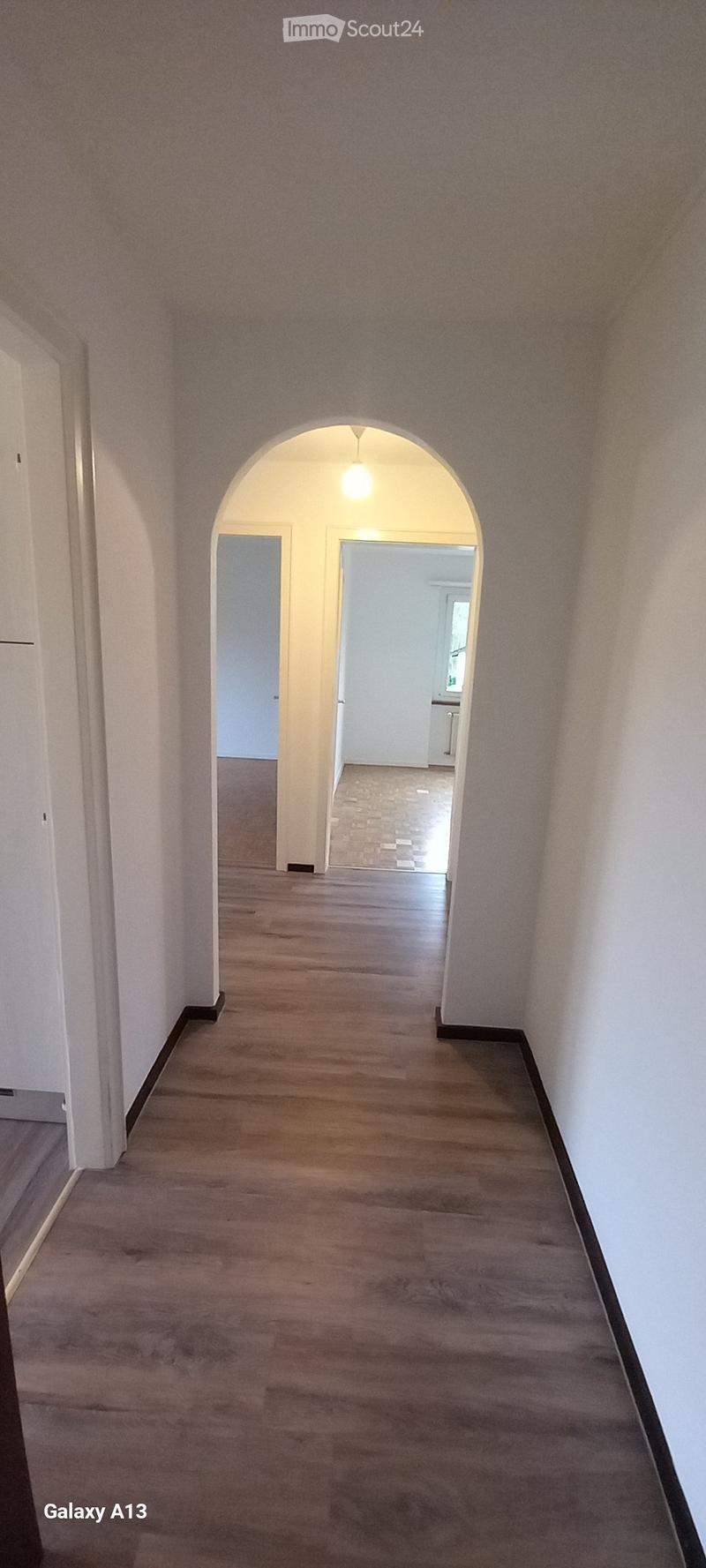 Empty hallway with wooden flooring, white walls, and an arched entrance. Two doors lead to adjacent rooms, and a bathroom with a glass door is visible.