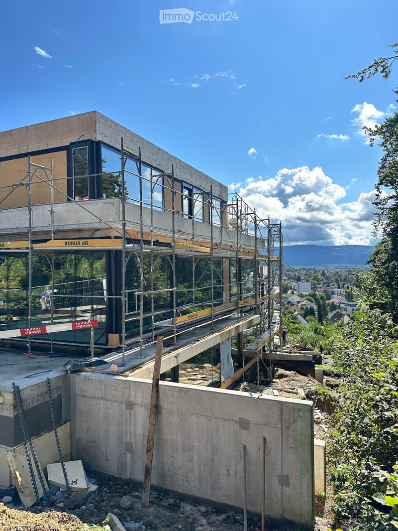 Modern house under construction with large windows, currently on second floor with scaffolding, surrounded by trees and mountains
