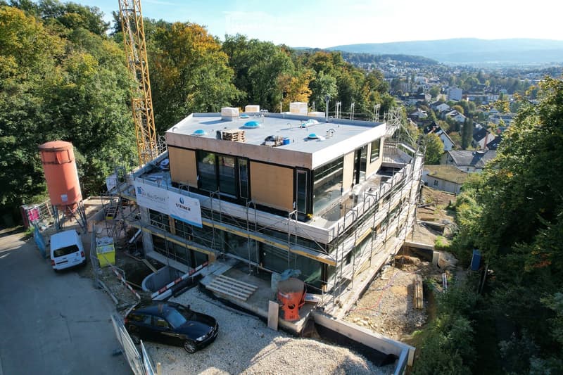 Aerial view of a new construction project, modern architecture with large glass windows, under construction with visible scaffolding and equipment on site, a crane adjacent to the building, a hilly terrain with visible vegetation.